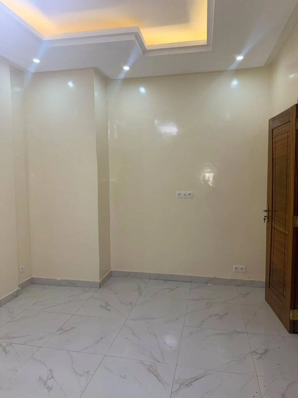 Pièce non meublée avec carrelage clair, porte en bois et éclairage indirect au plafond
